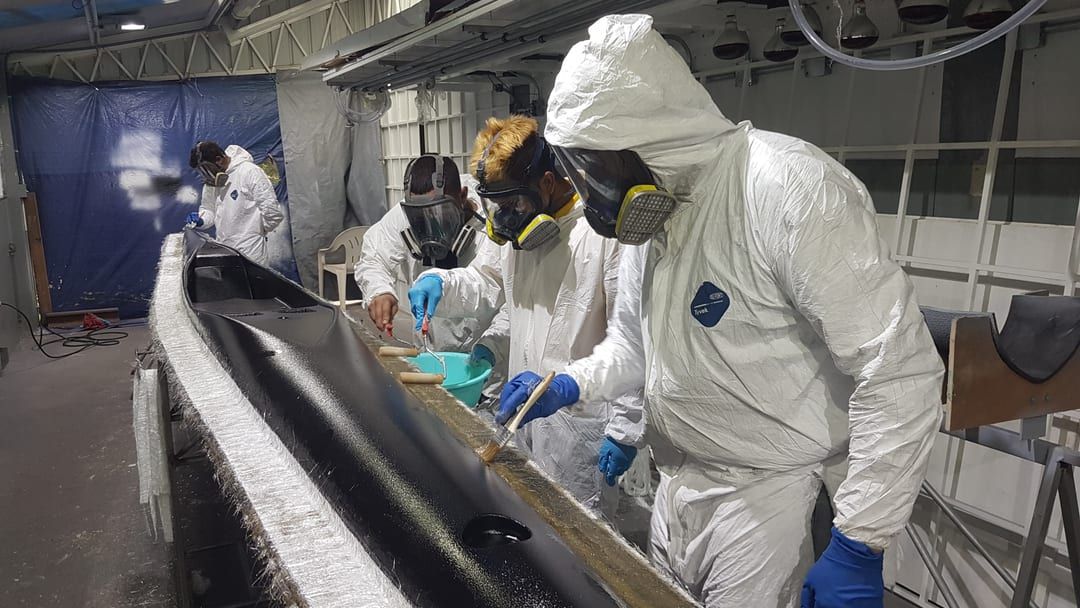 Tres personas con trajes blancos y respiradores de careta completa laminan un casco negro dentro de la cabina de pintura del taller.